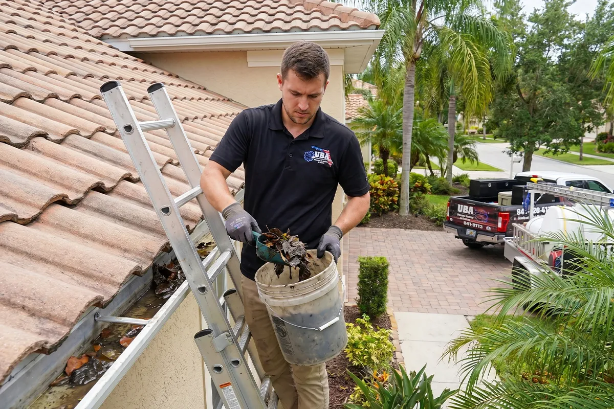 UBA technician on ladder removing gutter debris into bucket at a Sarasota tile roof home, equipment trailer visible in driveway