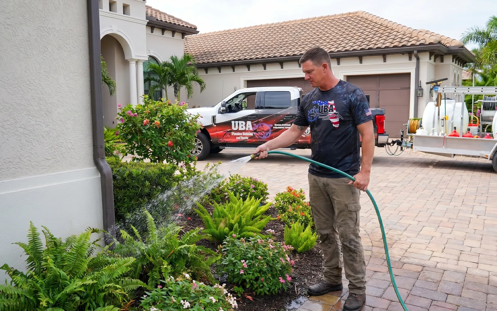 UBA Pressure Washing technician pre-wetting ornamental landscaping with a garden hose before soft wash application, protecting plants from chemical runoff, with UBA branded truck and tank trailer visible in the driveway