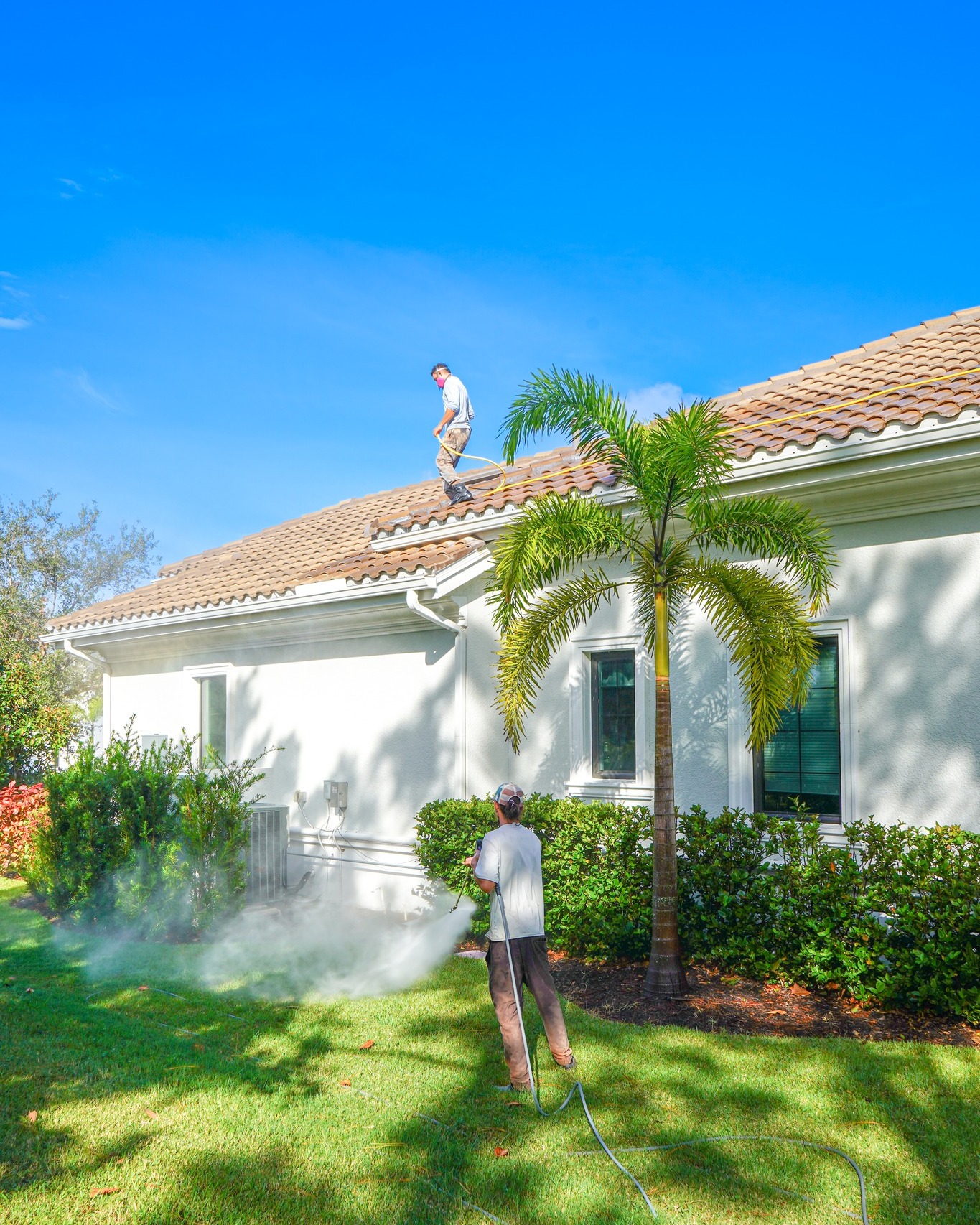 Two UBA Pressure Washing technicians working on a Sarasota barrel tile roof under blue Florida skies: one tech on the roof applying solution, one below at the ladder, with clean white home exterior visible